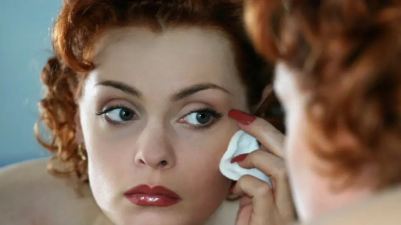 Donna dai capelli rossi si strucca il viso con un dischetto di cotone, guardandosi allo specchio.