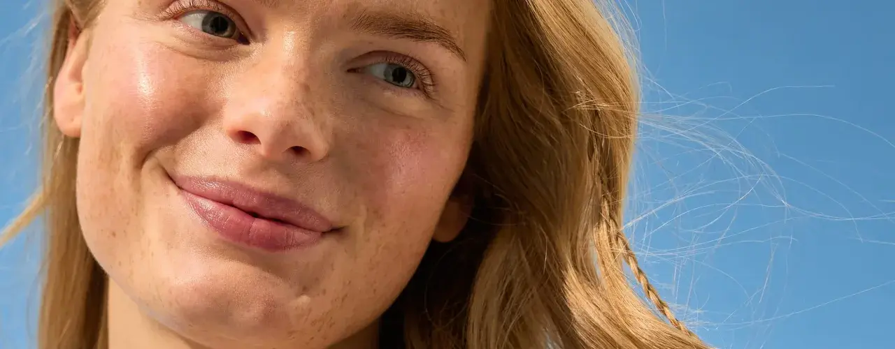 Viso sorridente con lentiggini, capelli biondi mossi dal vento. La sua pelle è protetta, una protezione 50 che dura 50 minuti sotto il sole.
