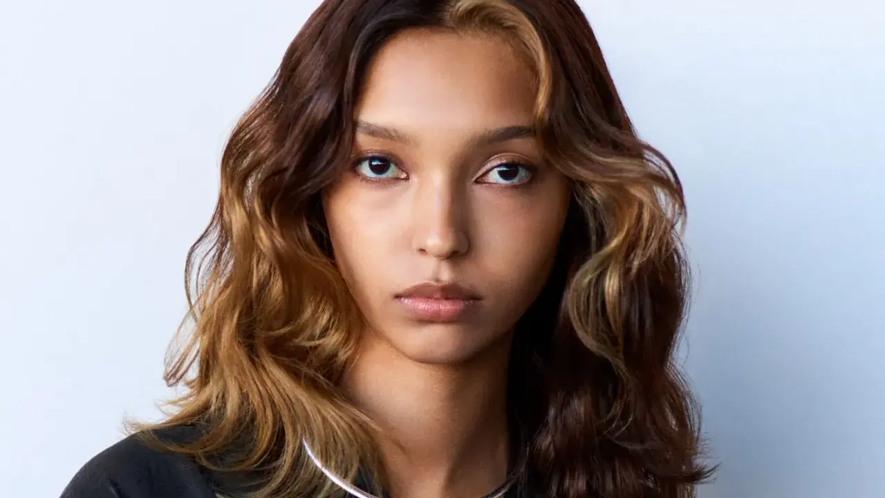 Volto di donna con capelli mossi castani e ciocche bionde, effetto colore demipermanente. Indossa una collana argentata.