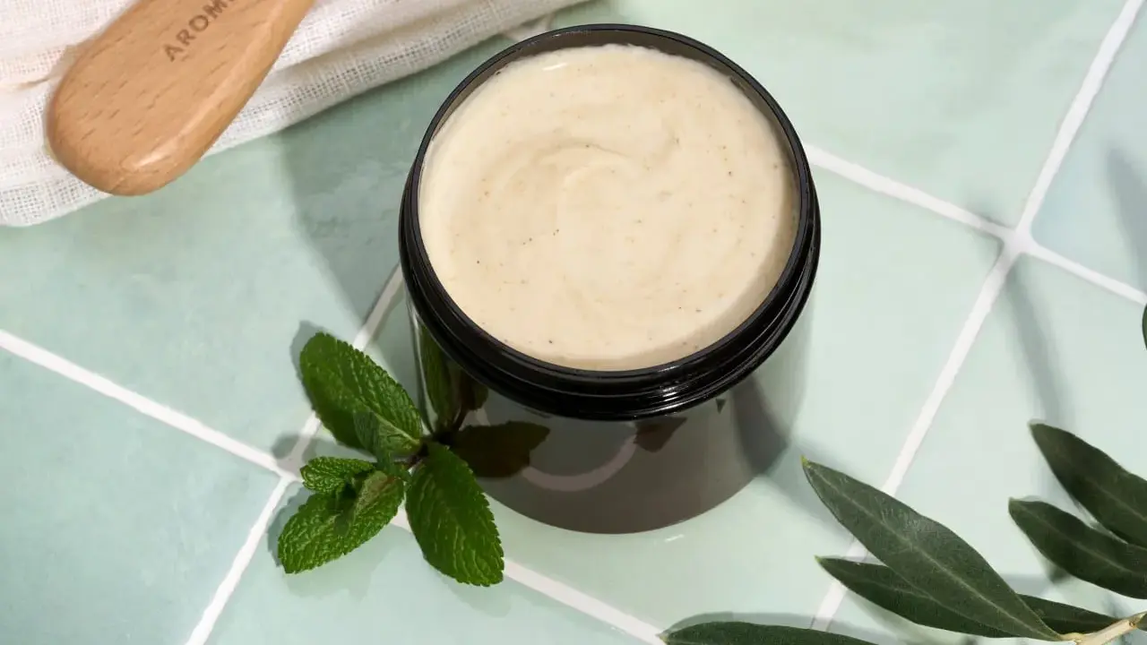 Vaso di scrub cuoio capelluto fai da te con menta e foglie d'ulivo. Un pettine in legno è visibile in alto a sinistra.