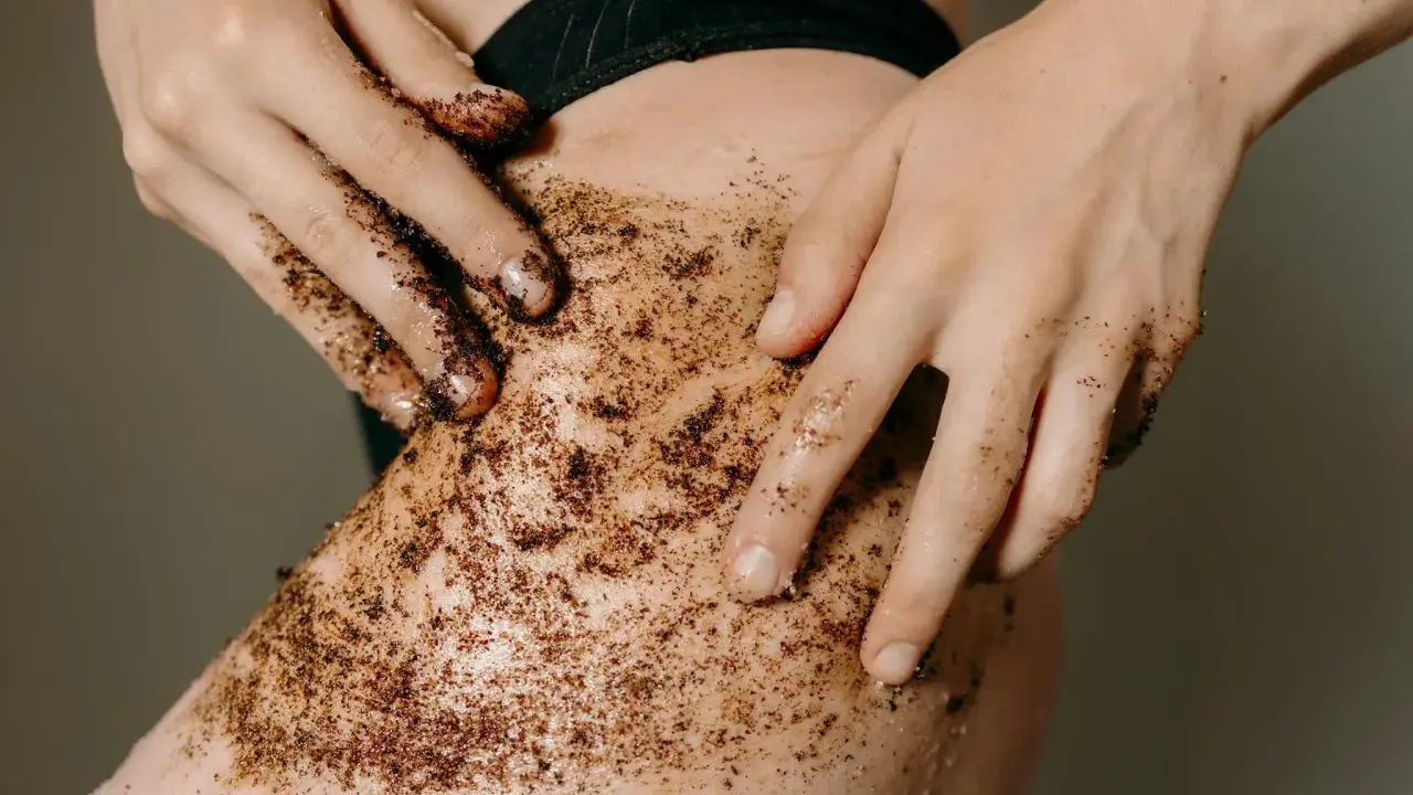 Mani che applicano uno scrub corpo al caffè sulla pelle. Un gesto per scoprire come fare lo scrub corpo e ottenere una pelle liscia e luminosa.