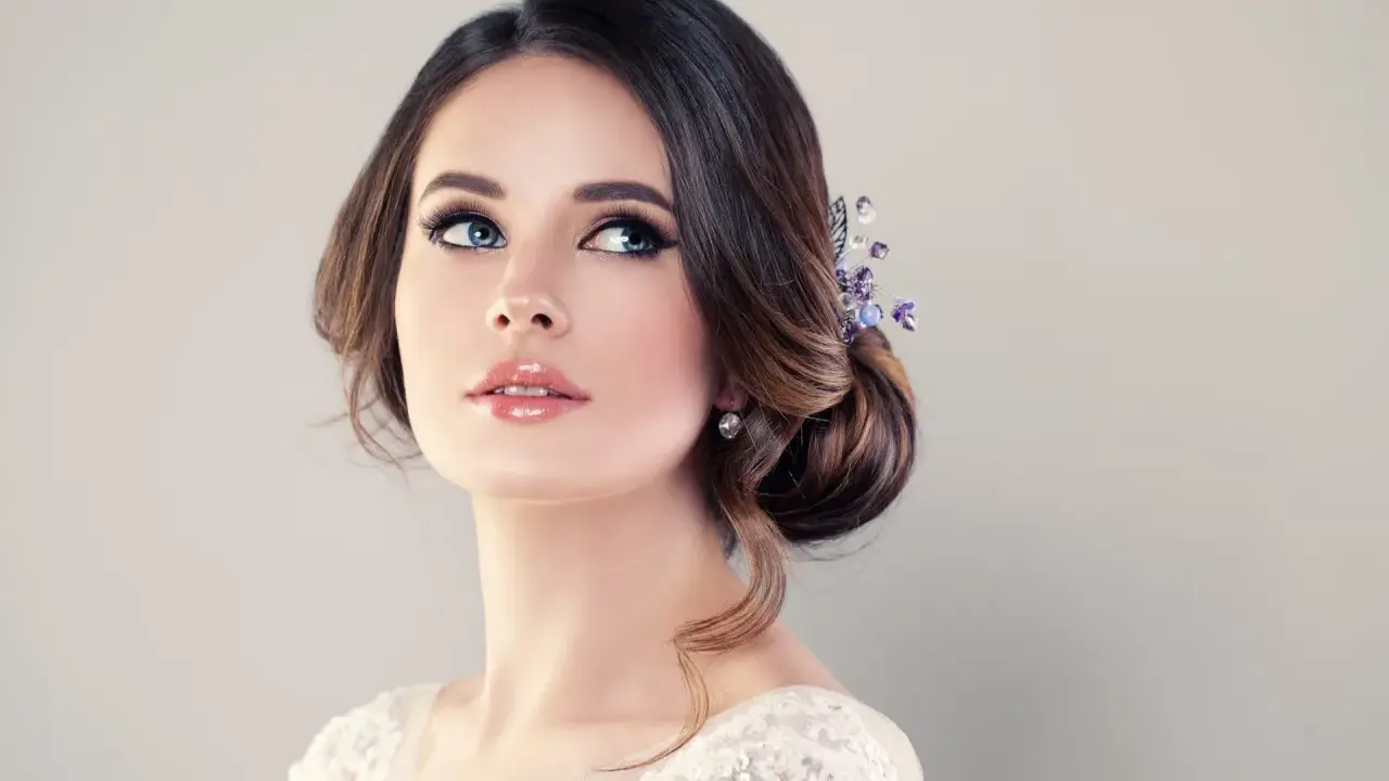 Donna con acconciatura raccolta e gioielli, labbra lucide e occhi azzurri intensi. Un trucco luminoso che esalta la sua bellezza naturale.