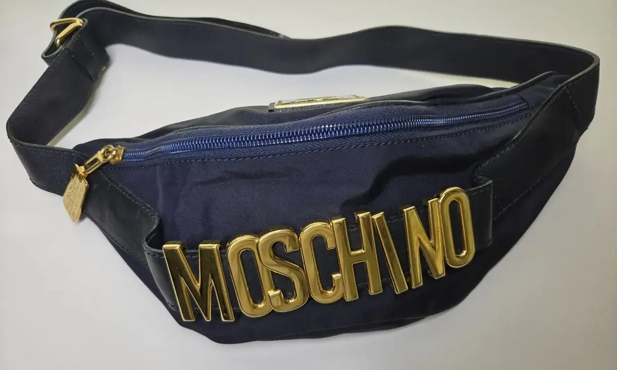 Borsa blu Moschino con logo dorato in rilievo, tracolla regolabile e zip. Un accessorio iconico per un look audace.