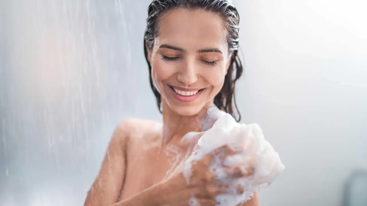 Donna sorridente sotto la doccia, insaponandosi con una spugna. Attenzione: il cocamidopropyl betaine fa male.