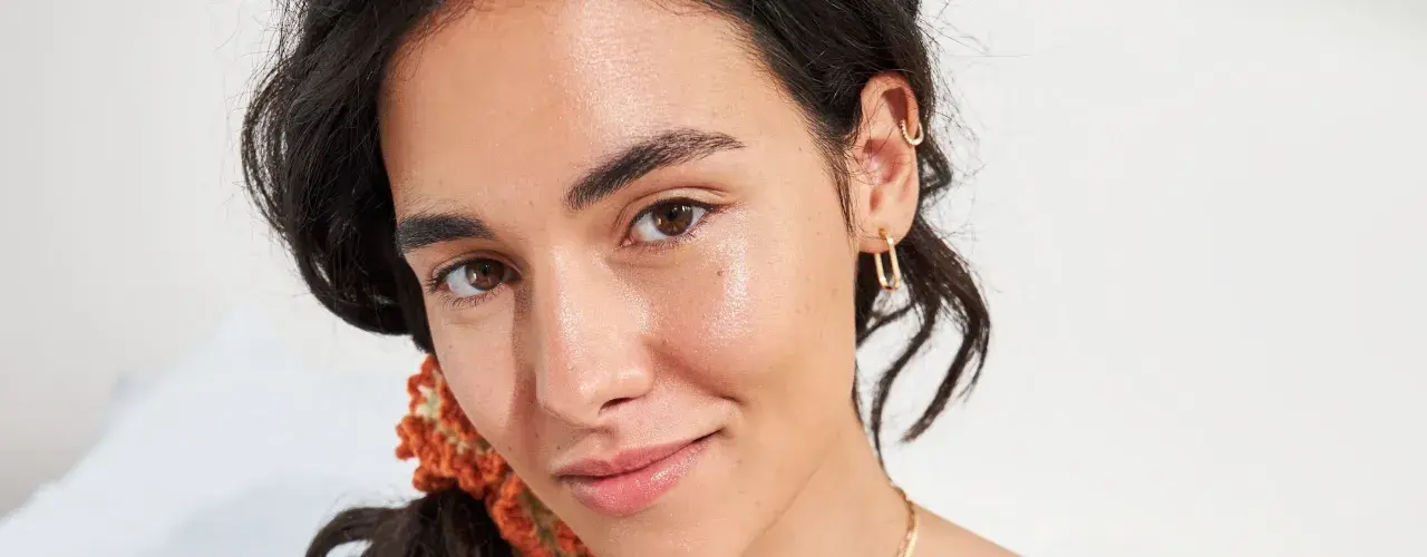Viso luminoso e idratato, con gocce di squalano che riflettono la luce. Gioielli dorati e capelli scuri completano il look.