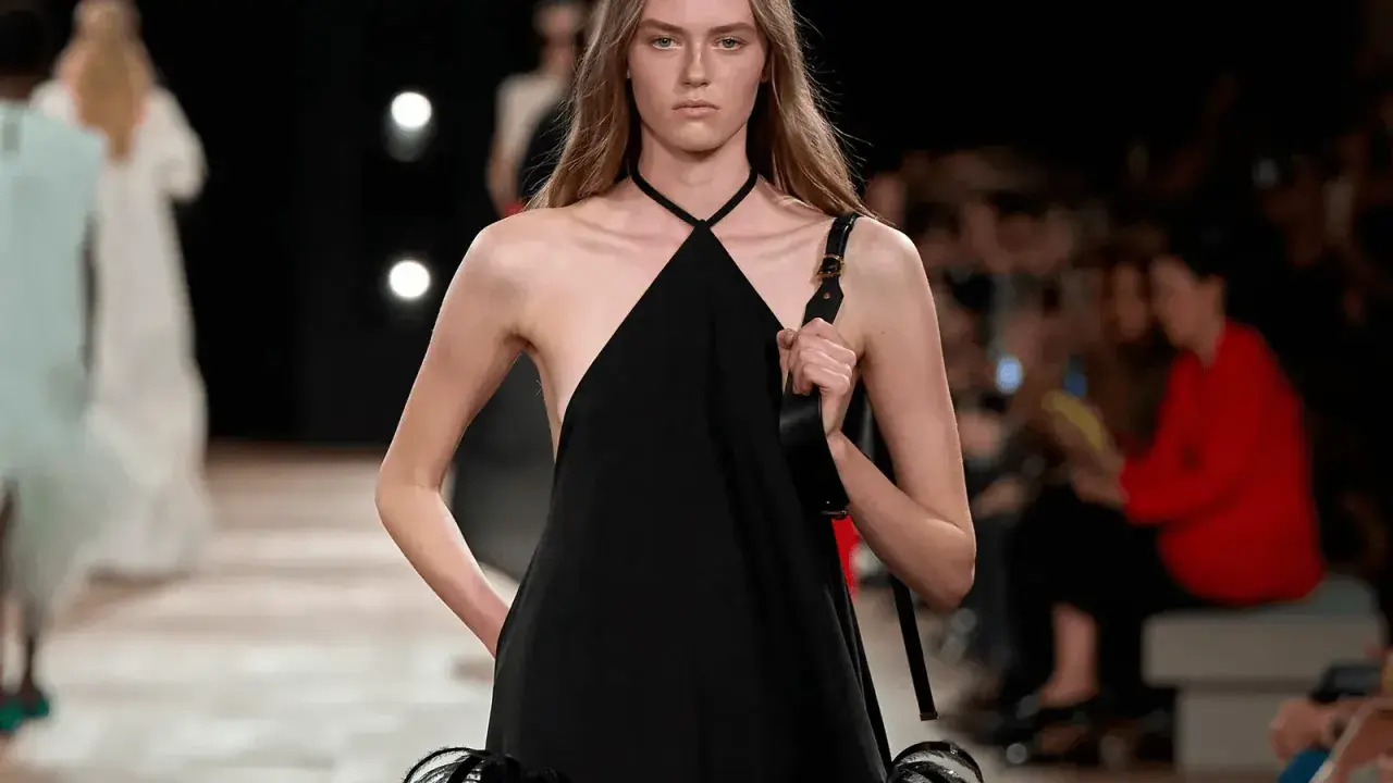Modella con un abito nero legato al collo, con dettagli di piume sulla gonna, sfila in passerella.