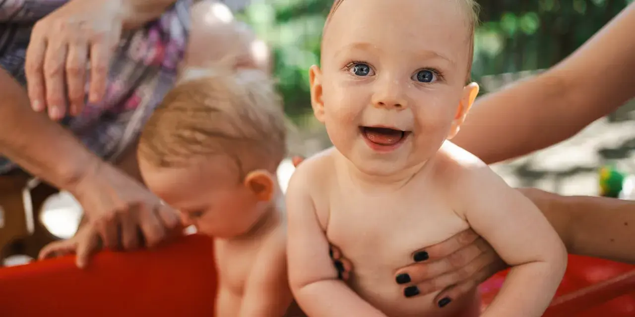 Bambini felici in una vasca rossa, con mani adulte che li sorreggono. Ricorda che la crema solare va messa sempre per proteggere la loro pelle delicata.