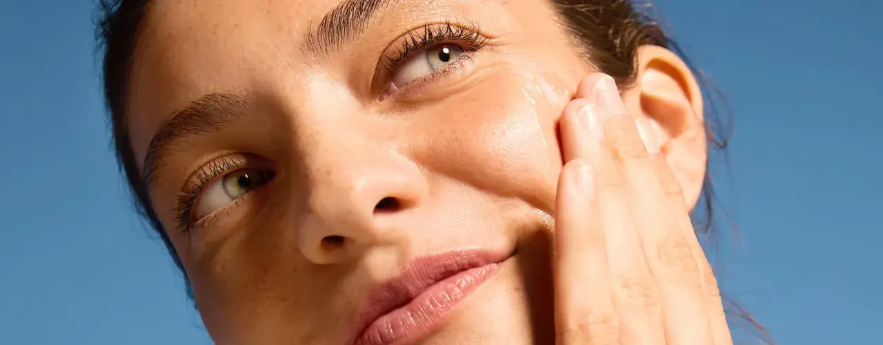 Primo piano di un volto femminile con pelle luminosa, mentre una mano applica delicatamente un prodotto sul viso.