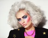 Icona pop con trucco anni 80 semplice: occhi blu elettrico, labbra rosa, capelli voluminosi e giacca nera con stelle dorate.
