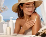 Donna con cappello di paglia applica crema solare sul braccio, mostrando che si può mettere il fondotinta sopra la crema solare per un look radioso.
