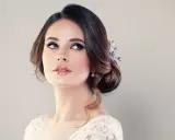 Donna con acconciatura raccolta e gioielli, labbra lucide e occhi azzurri intensi. Un trucco luminoso che esalta la sua bellezza naturale.