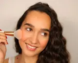 Donna sorridente applica il blush sulle guance con un pennello morbido, creando un effetto naturale e radioso.