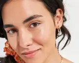 Viso luminoso e idratato, con gocce di squalano che riflettono la luce. Gioielli dorati e capelli scuri completano il look.