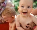 Bambini felici in una vasca rossa, con mani adulte che li sorreggono. Ricorda che la crema solare va messa sempre per proteggere la loro pelle delicata.