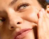 Primo piano di un volto femminile con pelle luminosa, mentre una mano applica delicatamente un prodotto sul viso.