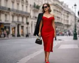 Donna elegante con un **abito rosso raso** scollato, cammina per strada con un blazer nero sulla spalla e una borsa.