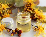 Un vasetto di olio dorato circondato da fiori gialli e boccioli rossi. L'amamelide cos'è? Un rimedio naturale.