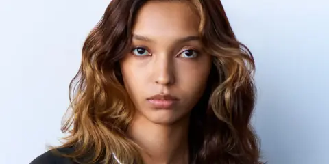 Volto di donna con capelli mossi castani e ciocche bionde, effetto colore demipermanente. Indossa una collana argentata.