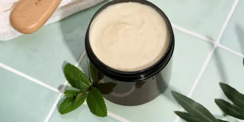 Vaso di scrub cuoio capelluto fai da te con menta e foglie d'ulivo. Un pettine in legno è visibile in alto a sinistra.