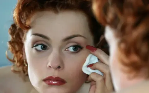 Donna dai capelli rossi si strucca il viso con un dischetto di cotone, guardandosi allo specchio.