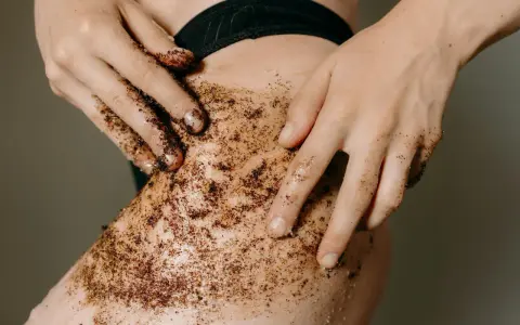 Mani che applicano uno scrub corpo al caffè sulla pelle. Un gesto per scoprire come fare lo scrub corpo e ottenere una pelle liscia e luminosa.