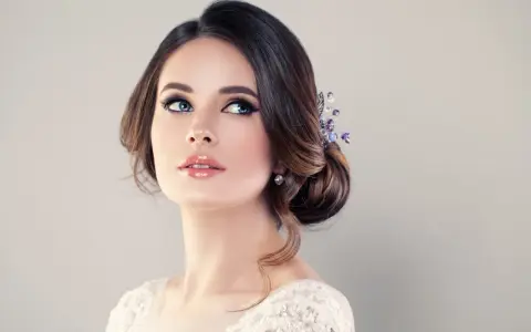 Donna con acconciatura raccolta e gioielli, labbra lucide e occhi azzurri intensi. Un trucco luminoso che esalta la sua bellezza naturale.