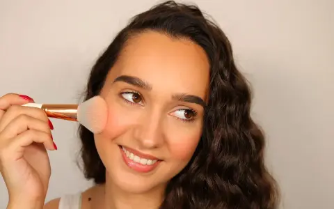 Donna sorridente applica il blush sulle guance con un pennello morbido, creando un effetto naturale e radioso.