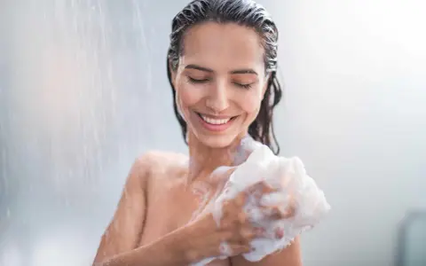 Donna sorridente sotto la doccia, insaponandosi con una spugna. Attenzione: il cocamidopropyl betaine fa male.