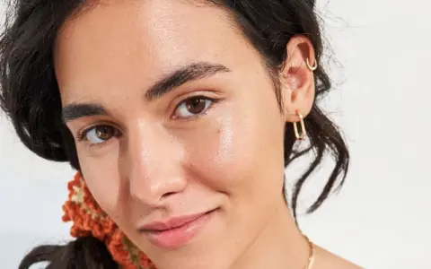 Viso luminoso e idratato, con gocce di squalano che riflettono la luce. Gioielli dorati e capelli scuri completano il look.