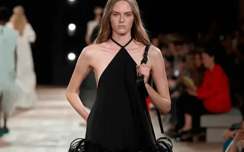 Modella con un abito nero legato al collo, con dettagli di piume sulla gonna, sfila in passerella.