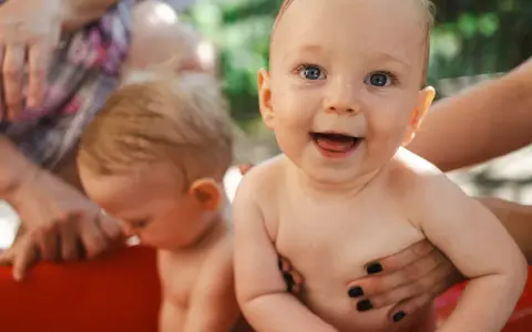 Bambini felici in una vasca rossa, con mani adulte che li sorreggono. Ricorda che la crema solare va messa sempre per proteggere la loro pelle delicata.