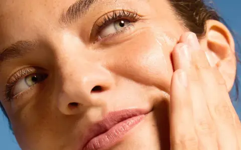 Primo piano di un volto femminile con pelle luminosa, mentre una mano applica delicatamente un prodotto sul viso.