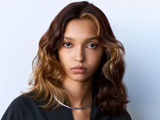 Volto di donna con capelli mossi castani e ciocche bionde, effetto colore demipermanente. Indossa una collana argentata.