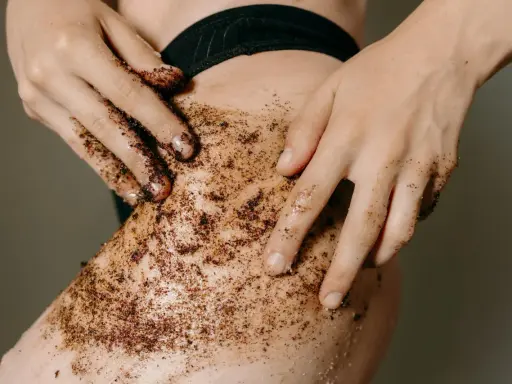 Mani che applicano uno scrub corpo al caffè sulla pelle. Un gesto per scoprire come fare lo scrub corpo e ottenere una pelle liscia e luminosa.