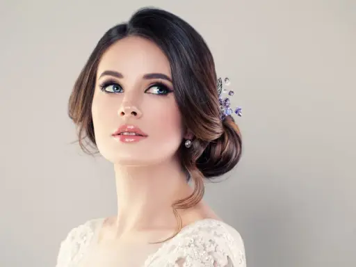 Donna con acconciatura raccolta e gioielli, labbra lucide e occhi azzurri intensi. Un trucco luminoso che esalta la sua bellezza naturale.