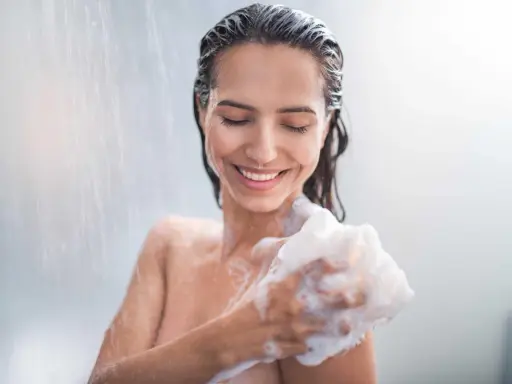 Donna sorridente sotto la doccia, insaponandosi con una spugna. Attenzione: il cocamidopropyl betaine fa male.