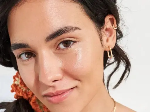 Viso luminoso e idratato, con gocce di squalano che riflettono la luce. Gioielli dorati e capelli scuri completano il look.