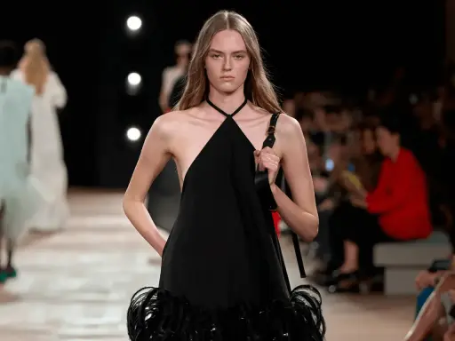 Modella con un abito nero legato al collo, con dettagli di piume sulla gonna, sfila in passerella.