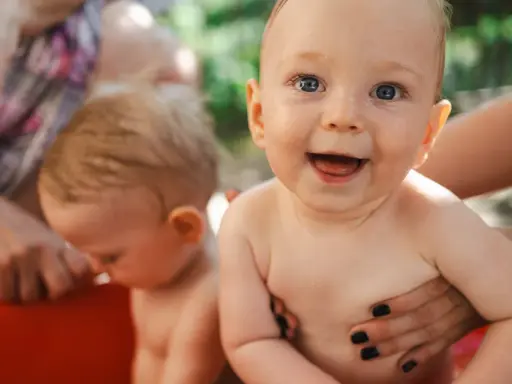 Bambini felici in una vasca rossa, con mani adulte che li sorreggono. Ricorda che la crema solare va messa sempre per proteggere la loro pelle delicata.