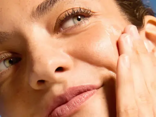 Primo piano di un volto femminile con pelle luminosa, mentre una mano applica delicatamente un prodotto sul viso.