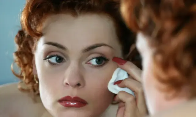 Donna dai capelli rossi si strucca il viso con un dischetto di cotone, guardandosi allo specchio.