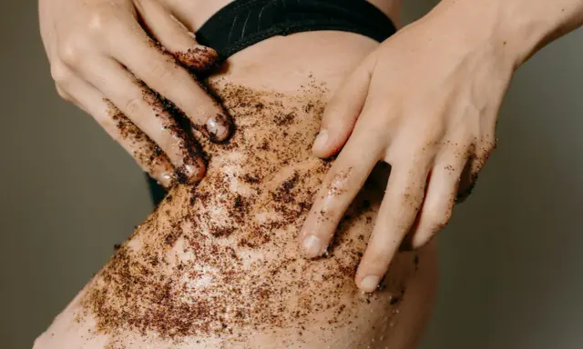 Mani che applicano uno scrub corpo al caffè sulla pelle. Un gesto per scoprire come fare lo scrub corpo e ottenere una pelle liscia e luminosa.