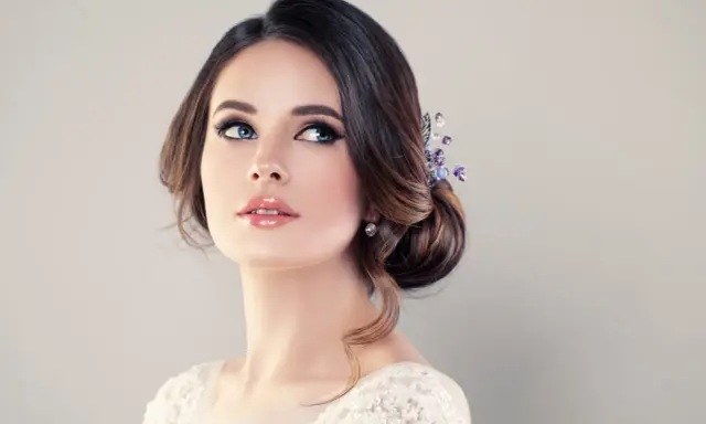 Donna con acconciatura raccolta e gioielli, labbra lucide e occhi azzurri intensi. Un trucco luminoso che esalta la sua bellezza naturale.