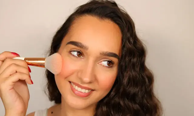 Donna sorridente applica il blush sulle guance con un pennello morbido, creando un effetto naturale e radioso.