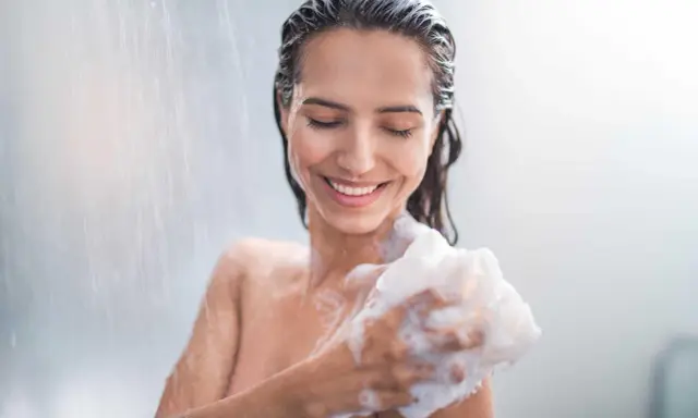 Donna sorridente sotto la doccia, insaponandosi con una spugna. Attenzione: il cocamidopropyl betaine fa male.