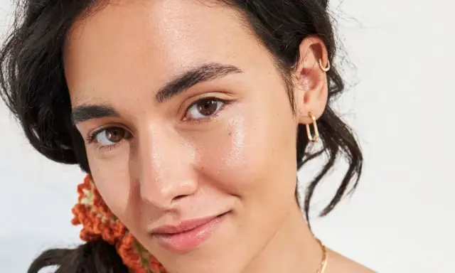 Viso luminoso e idratato, con gocce di squalano che riflettono la luce. Gioielli dorati e capelli scuri completano il look.