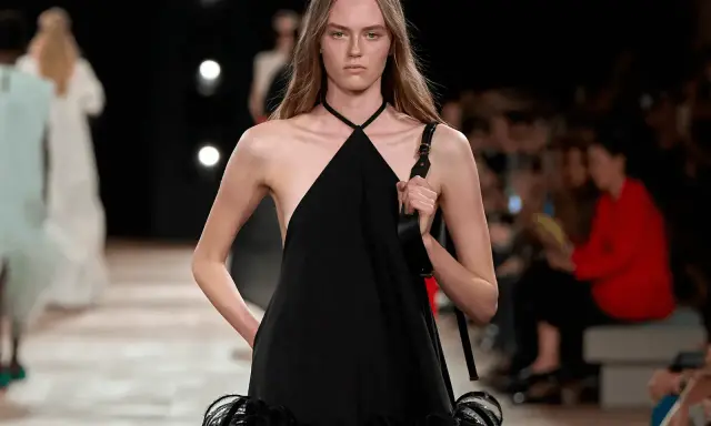 Modella con un abito nero legato al collo, con dettagli di piume sulla gonna, sfila in passerella.
