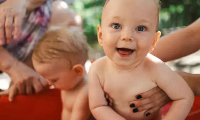 Bambini felici in una vasca rossa, con mani adulte che li sorreggono. Ricorda che la crema solare va messa sempre per proteggere la loro pelle delicata.