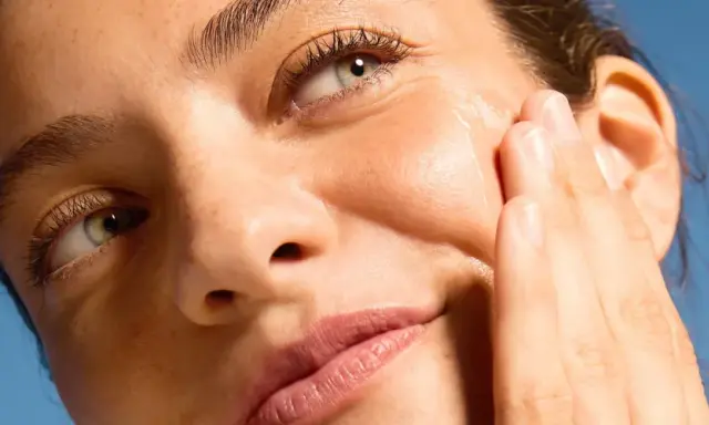 Primo piano di un volto femminile con pelle luminosa, mentre una mano applica delicatamente un prodotto sul viso.