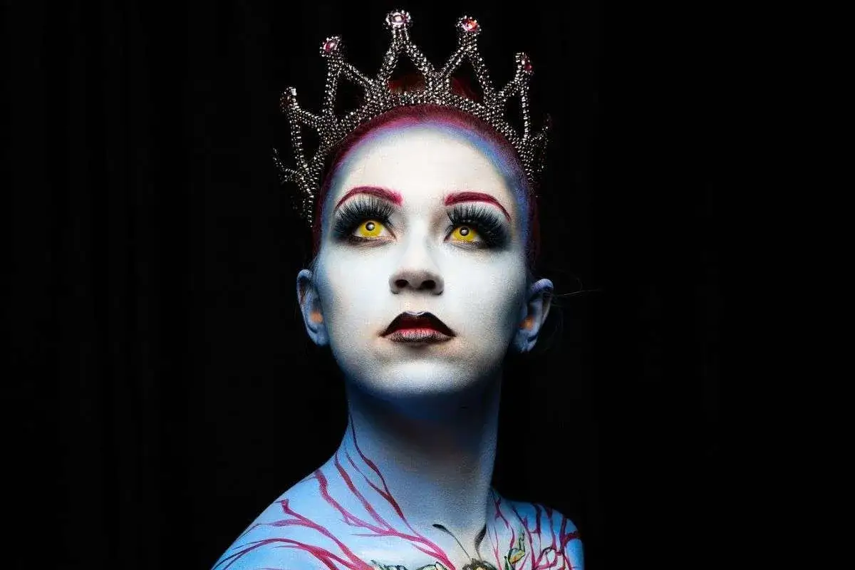 Regina con trucco artistico: pelle azzurra, occhi gialli, corona scintillante e decorazioni sul corpo. Idee trucco artistico per un look da favola oscura.